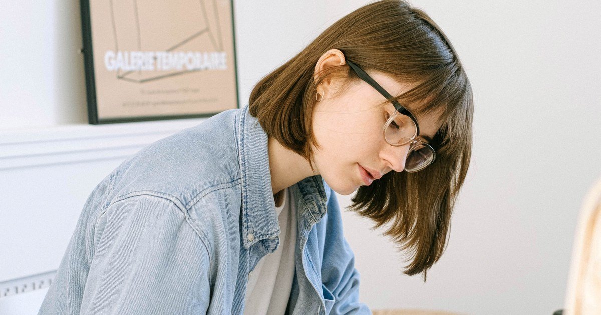 Junge Frau mit Kurzhaarschnitt und Brille in Jeansjacke, konzentriert nach unten blickend, im Hintergrund gerahmtes Poster „Galerie Temporaire“.