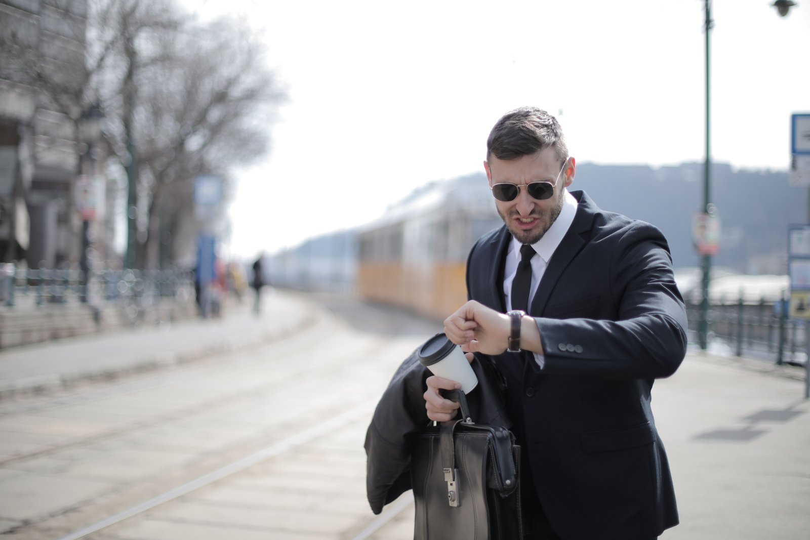 Geschäftsmann in Anzug mit Sonnenbrille und Koffer am Bahnhof, schaut auf Uhr.