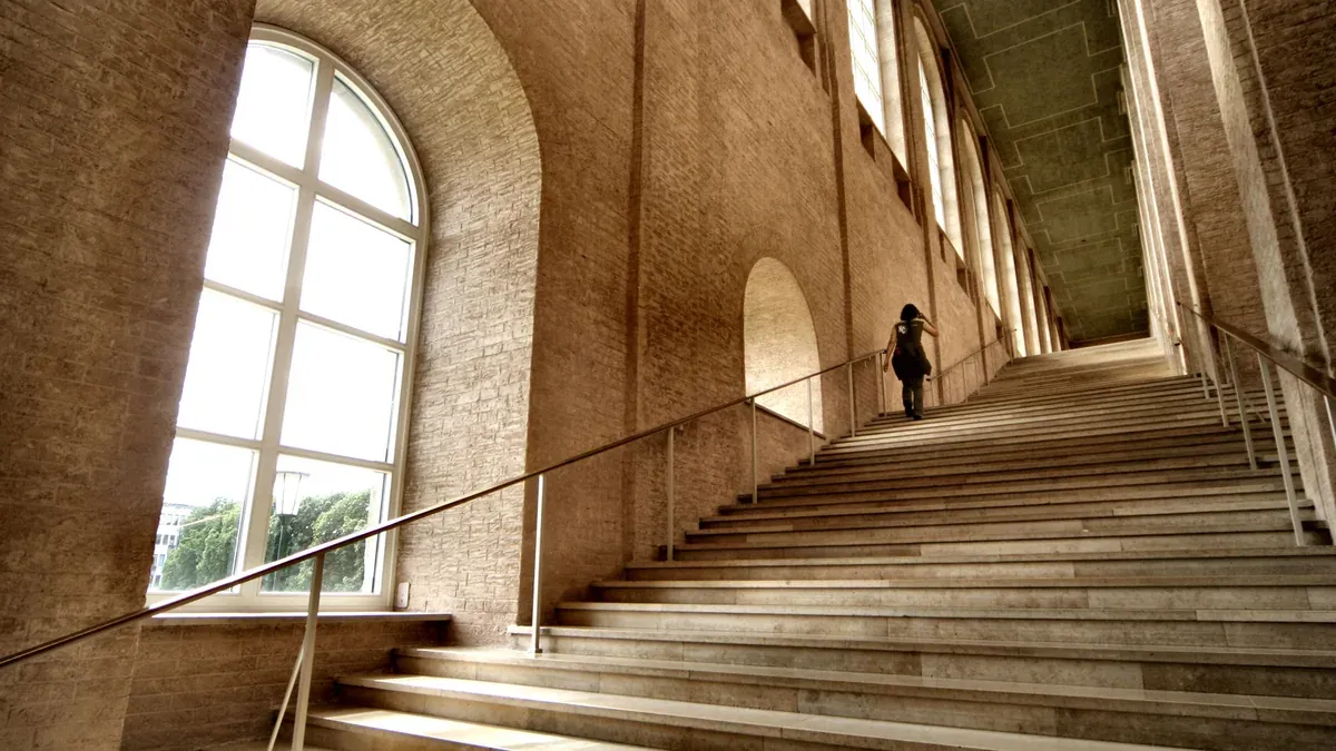 Person geht große Steintreppe hinauf in historischem Gebäude mit Bogenfenstern und Backsteinwänden