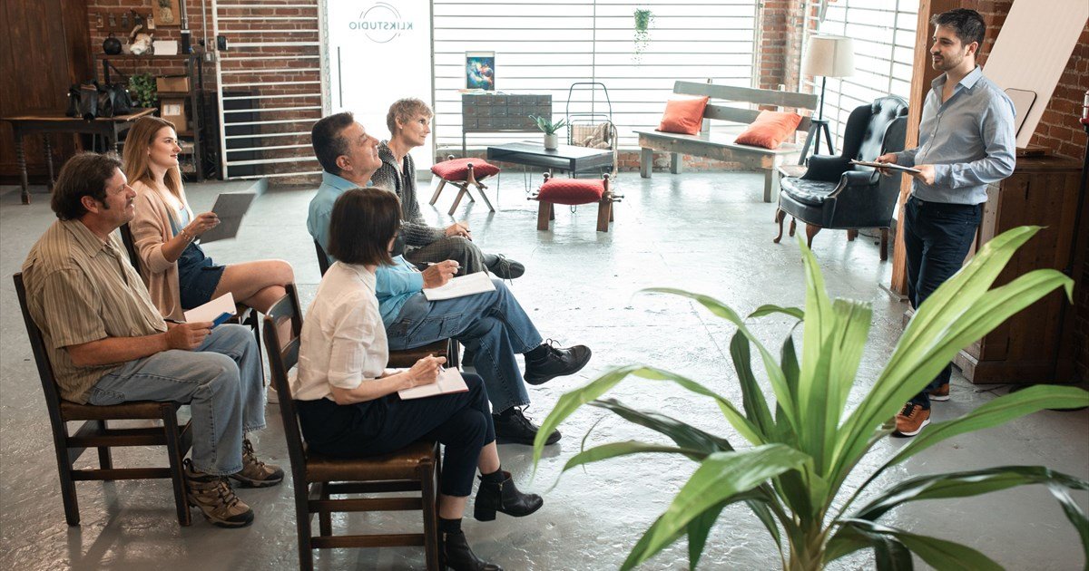 Gruppe sitzt im Halbkreis auf Stühlen und hört einem Mann zu, der in einem modernen Loft-Studio eine Präsentation hält.