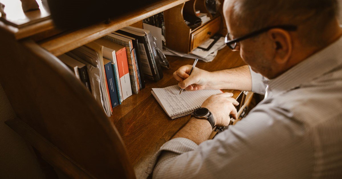 Mann mit Brille schreibt handschriftliche Notizen in Notizbuch am Holzschreibtisch mit Büchern im Hintergrund