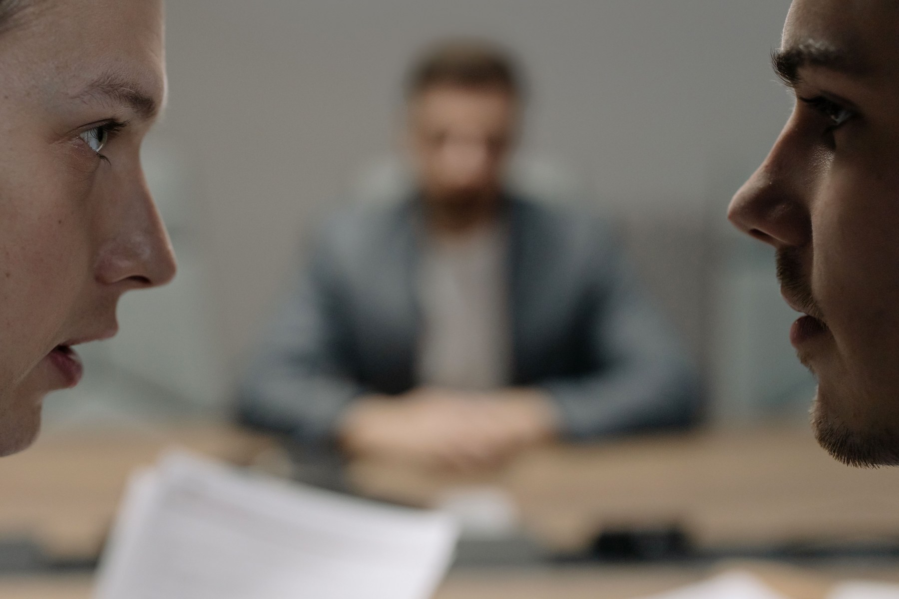Zwei Männer stehen sich in einer geschäftlichen Situation ernst gegenüber; eine dritte Person sitzt unscharf dahinter.