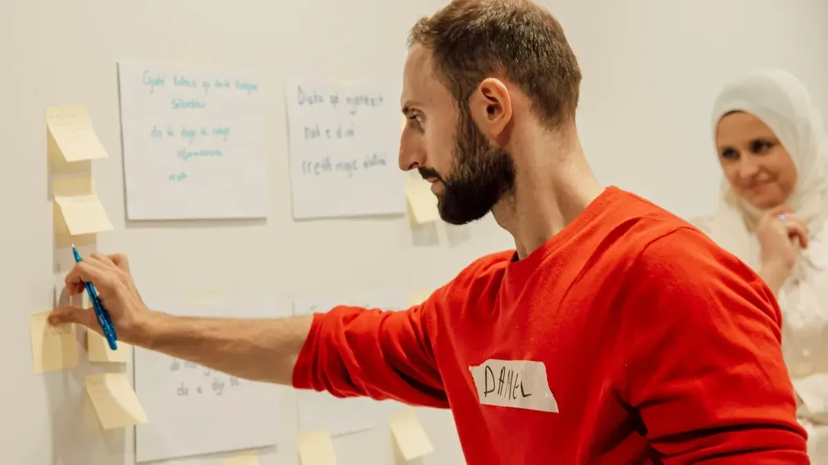 Mann klebt Haftnotizen an eine Wand, im Hintergrund eine lächelnde Teilnehmerin