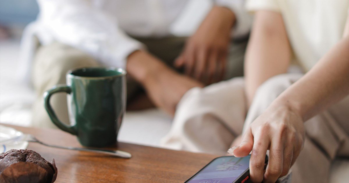Person entspannt mit Smartphone am Holztisch, grüne Kaffeetasse und Schokomuffin, gemütliche Wohnzimmerszene