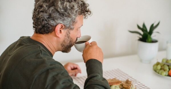 Mann mit grauen Locken trinkt Kaffee am Frühstückstisch mit Sandwich, Trauben und Pflanze im Hintergrund