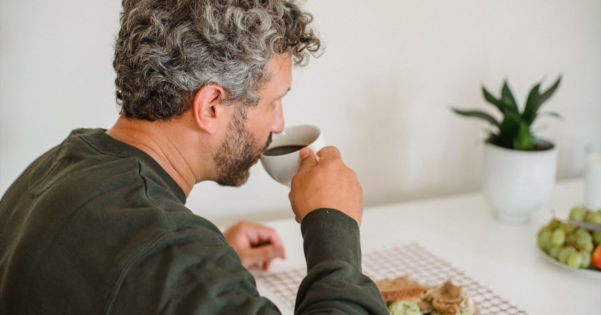 Mann mit grauen Locken trinkt Kaffee am Frühstückstisch mit Sandwich, Trauben und Pflanze im Hintergrund