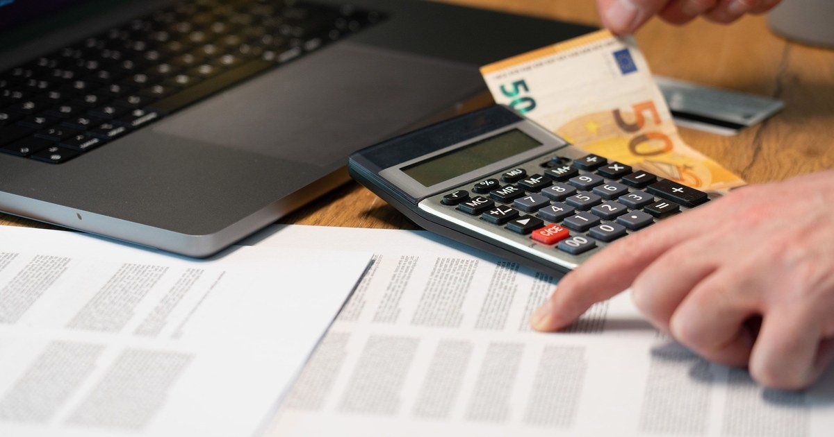 Person berechnet Finanzen mit Taschenrechner, Euro-Geldschein, Laptop und Dokumenten auf Schreibtisch