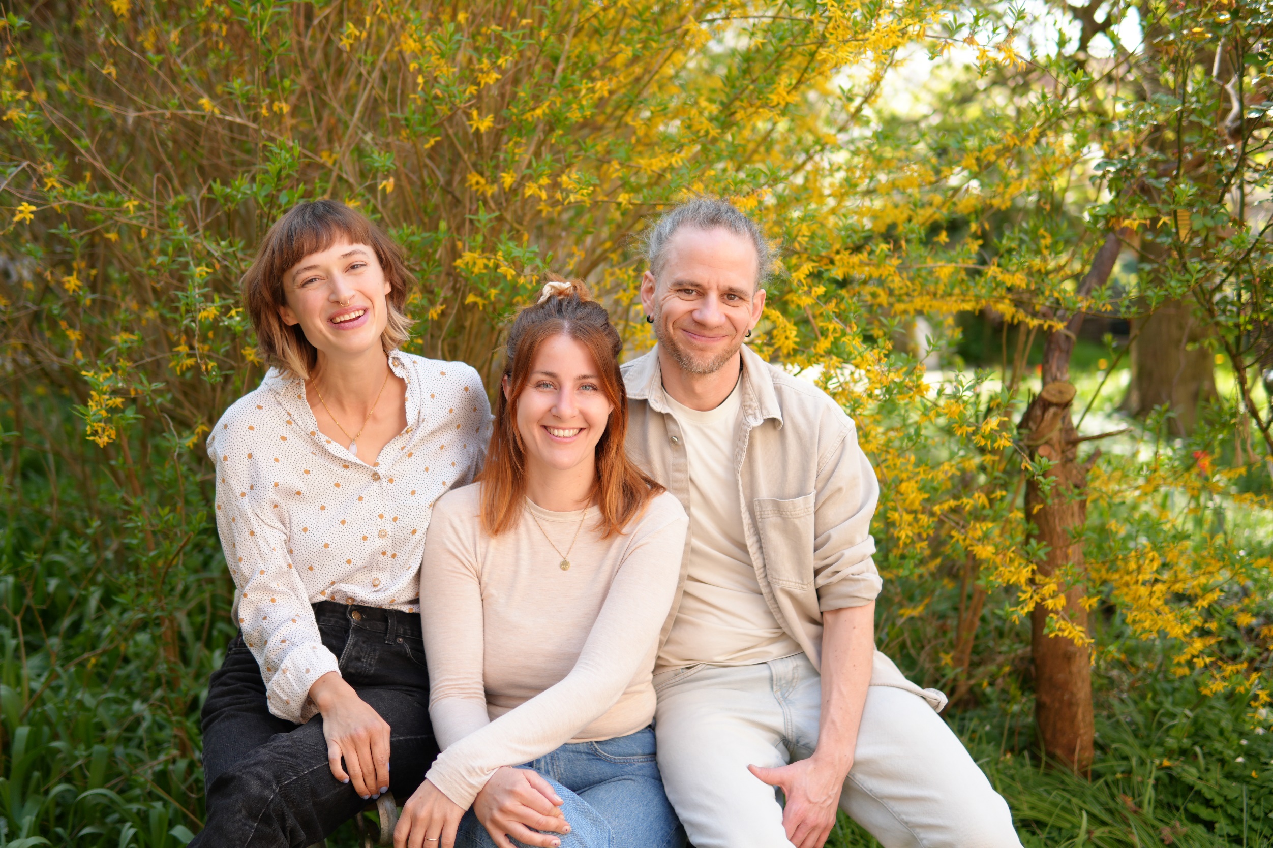 Ronja Junicke, Teresa Hierenbach und Markus Castro sitzen nebeneinander vor einem blühenden Forsythienstrauch