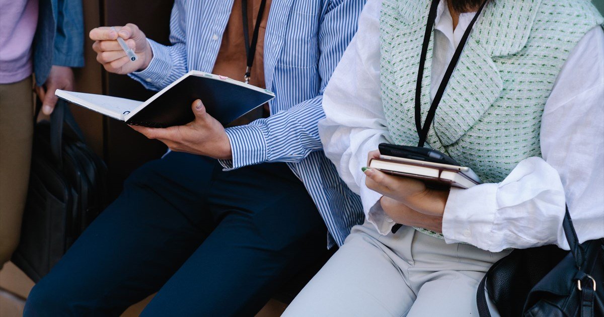 Zwei Personen sitzen mit Notizbüchern und Stift, tragen Lanyards, im Business-Casual-Look bei einem Meeting.