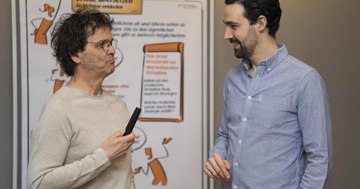 Zwei Trainer im Gespräch vor einem Flipchart mit der Überschrift Urteile übersetzen, Bedürfnisse entdecken.