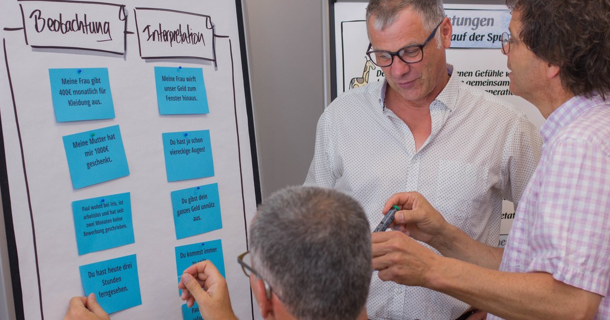 Drei Menschen stehen im Seminarraum zusammen und besprechen Karten, im Hintergrund ein Flipchart.