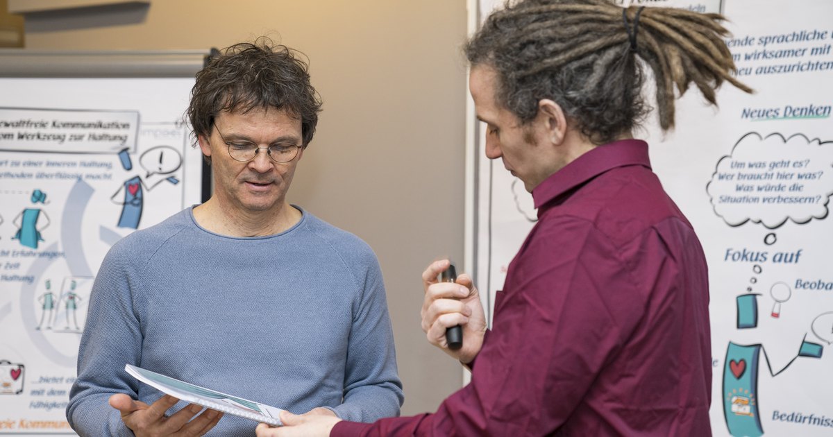Zwei Männer im Gespräch vor einem Flipchart, einer hält ein Blatt und hört zu.