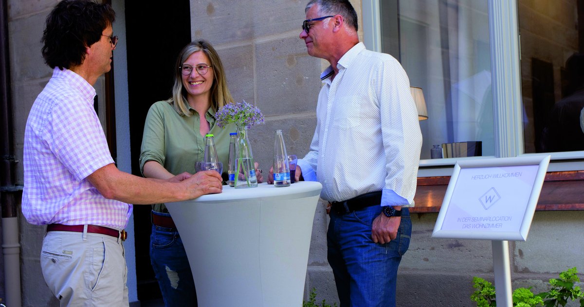 Drei Menschen stehen an einem Stehtisch im Innenhof des Seminarhauses und sprechen miteinander.
