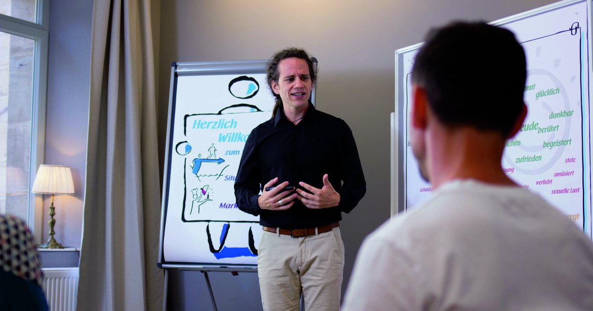 Ein Trainer steht vor einem Flipchart mit der Überschrift Herzlich willkommen und spricht zu Teilnehmenden.