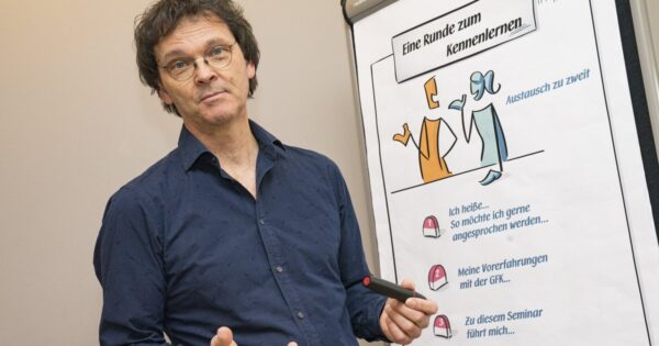 Ein Trainer erklärt vor einem gezeichneten Flipchart mit der Überschrift Eine Runde zum Kennenlernen.
