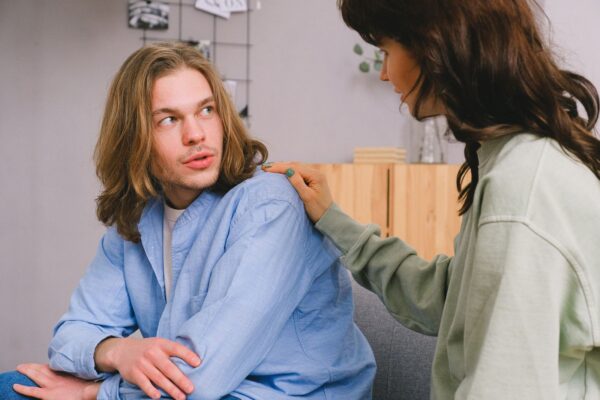 Frau legt tröstend die Hand auf die Schulter eines nachdenklichen Mannes mit langen Haaren im Gespräch