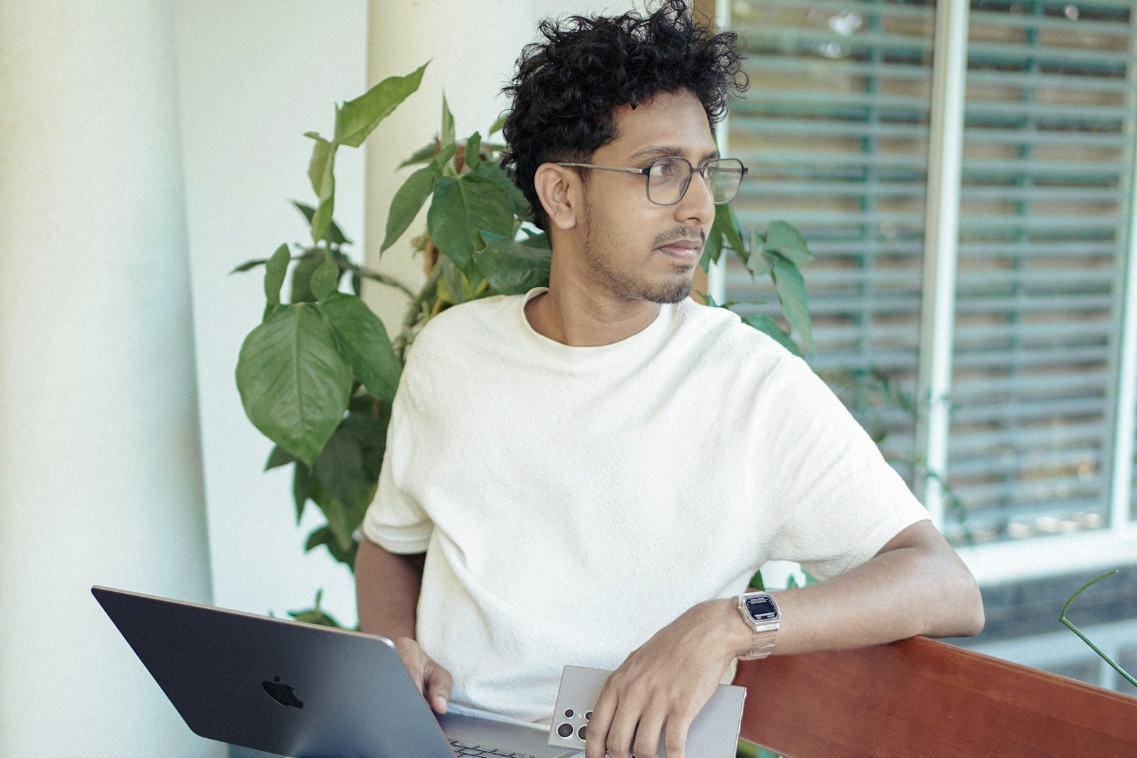 Mann mit Locken und Brille sitzt mit MacBook und Smartphone am Tisch, blickt nachdenklich zur Seite, im Hintergrund grüne Pflanze.