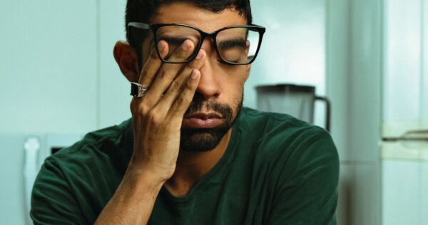 Mann mit Brille sitzt am Küchentisch und reibt sich mit der Hand das Auge.